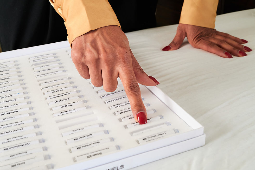 Hands with red nails point to labeled applied kinesiology vials on a white tray used for muscle testing. The person wears a beige shirt. Vials have various labels. Setting is neutral.