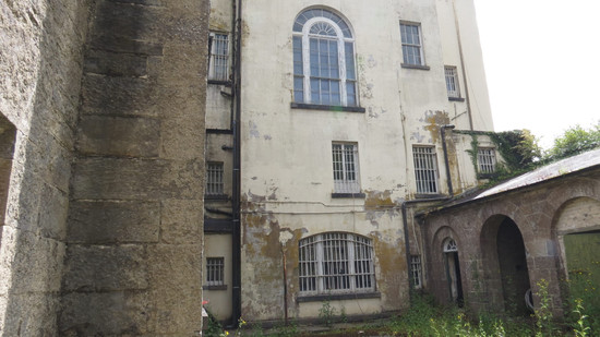 The side of a dilapidated country house - the paint is peeling, the ground is overgrown. There are a variety of windows on the facade, including a large arched window.