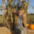 Colleen sits on a hay bail in a pumpkin patch where sunlight brightens the scene! She wears a yellow toque, rainbow tasseled earrings, large clear glasses, and a jacket over a hoodie.