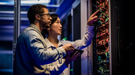 Dois analistas examinam um rack de servidores, concentrando-se na avaliação dos cabos e conexões em um ambiente de data center.
