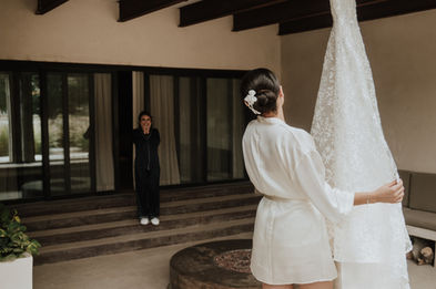 De espaldas, la novia sostiene un costado del vestido colgado mientras su mamá observa desde el fondo del corredor.