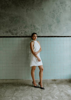 Emily posa con los brazos suavemente cruzados al frente, vestido blanco corto de cuello halter y tacones negros, junto a una