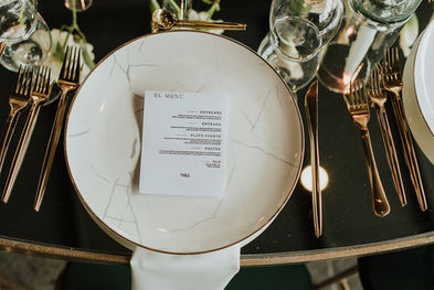 Detalle de la mesa con menú impreso, platos con borde dorado y cubiertos alineados con estética editorial.