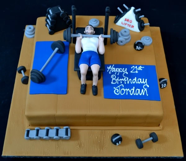 White fondant cake decorated with a gym-themed design featuring a weightlifter lifting a barbell on top of a square base.