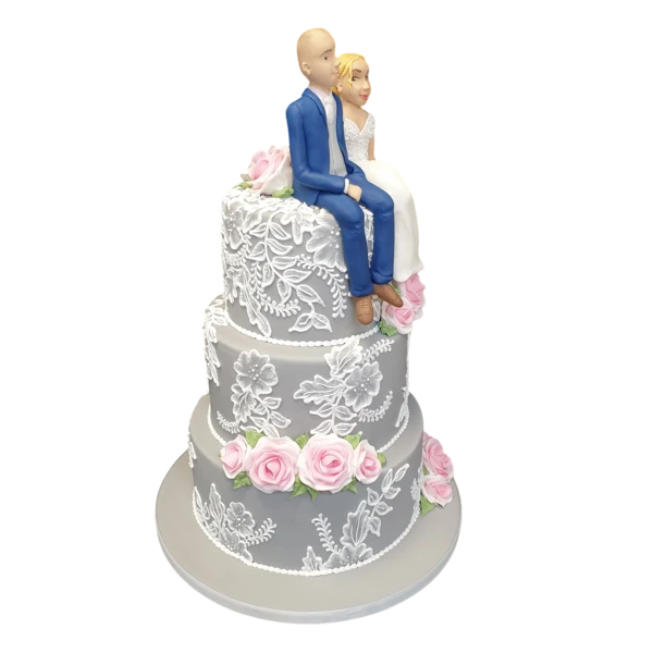 Three-tiered wedding cake with sugar paste lace and pink roses, topped with figurines.