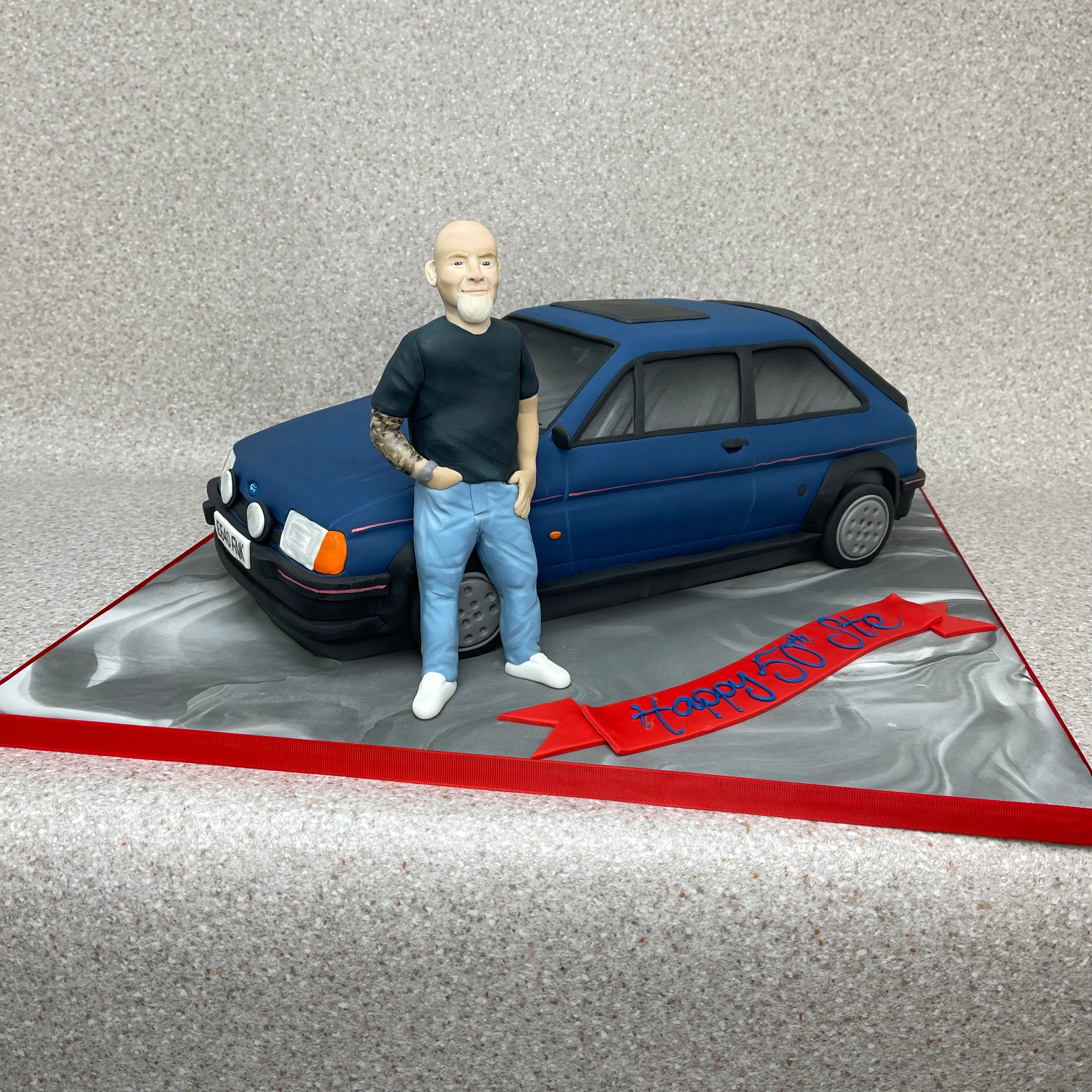 A retro-style celebration cake featuring a fondant figurine of a man standing next to a classic Fiesta XR2 car, creating a fun and unique design. Perfect for car enthusiasts and special occasions.