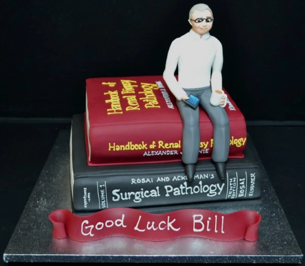 A unique cake inspired by a stack of pathology books with a figure on top, perfect for book lovers and special occasions.