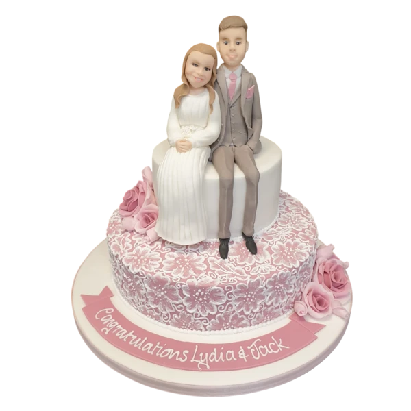 Two-tier wedding cake with sugar paste lace, roses, and personalised figurines.