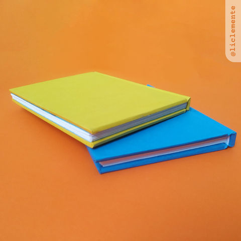 Na imagem vemos dois cadernos meio sobrepostos. O de baixo é azul claro e o de cima é amarelo. O de baixo só aparece metade da capa e o miolo branco entre as capas. No caderno de cima vemos toda a capa e o canto do caderno está virado para frente, assim vemos a lateral e a parte de baixo do miolo.