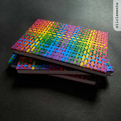 Foto de três cadernos empilhados, na qual a capa do caderno do topo é bem visível e nos de baixo apenas alguns cantos e suas lombadas de papel branco. A capa é formada por tiras de papel colorido entrelaçada formando um conjunto tecido colorido. 