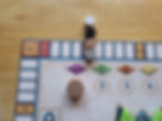 A close-up of the upper-left corner of the Summer Pavilion extra board. A scoring track runs around the outside perimeter, starting at 1 and running to 80 in the top-left corner. Two wooden cylinders sit on the 5 spot, and two more sit next to the 5 spot off the board because the 5 spot isn't big enough to hold all the scoring markers for all the players. The wooden cylinders are white, black, beige, and gray, to distinguish between the four players. Below the scoring track are the first four spaces of the round track. A large wooden cylinder sits on the one space to show that it is currently round 1. Above the 1 space is a purple diamond, above the 2 is a green diamond with a dark green geometric design, above the 3 is an orange diamond with a dark green geometric design, and above the 4 is a yellow diamond.