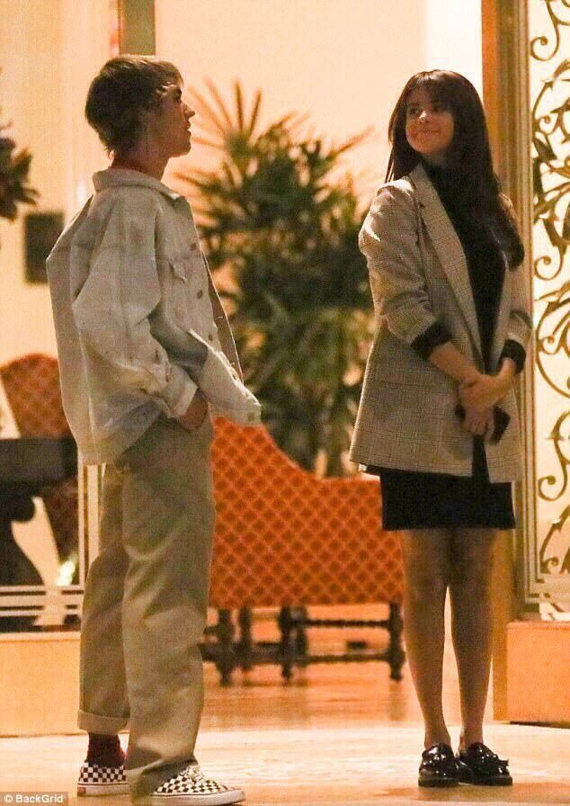 Deux personnes discutent dans un hall éclairé. L'un porte une veste en jean et des chaussures à carreaux, l'autre une robe noire avec un manteau.