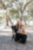 Sara, a lady with long blonde hair sits with her arm around a happy black and white border collie mix dog in a local park with lots of trees