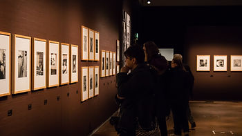 People viewing framed black and white photos in a dimly lit gallery. Dark walls and thoughtful expressions create a contemplative mood.