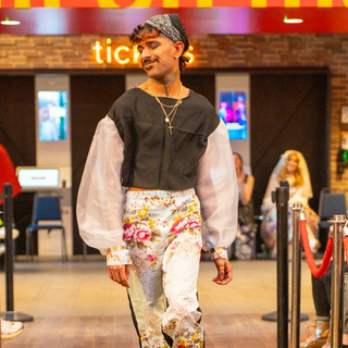 Person walking on catwalk wearing black and white bandana on head, black top with white billowing sleeves and  floral trousers on a light blue back ground.