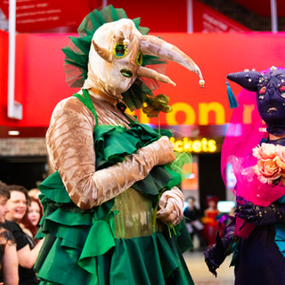 Performer in slug costume wearing beige leopard velvet and green chiffon dress with ruffles standing next to a sea slug wearing a black mask and magenta chiffon veil and pink blue and purple sash representing the bisexual flag with long black gloves holding a bunch of pink artificial roses.