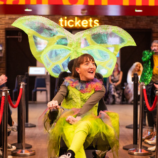 Woman in electric wheelchair wearing lime green chiffon dress with lime green and turquoise butterfly wings above head.