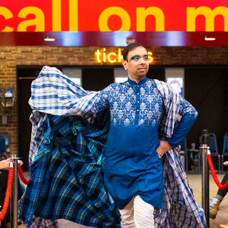 Male performer with blue eye make up walking on catwalk wearing blue Indian style long shirt with silver embroidery on and white trousers and bare feet. Blue and white chequered cape flowing behind.