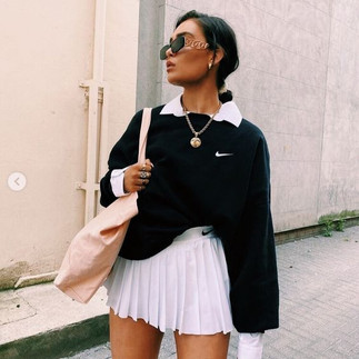 garota vestindo minissaia plissada branca esportiva da Nike, moletom preto da Nike, maxi acessórios e colar dourado, shopping bag e óculos escuros