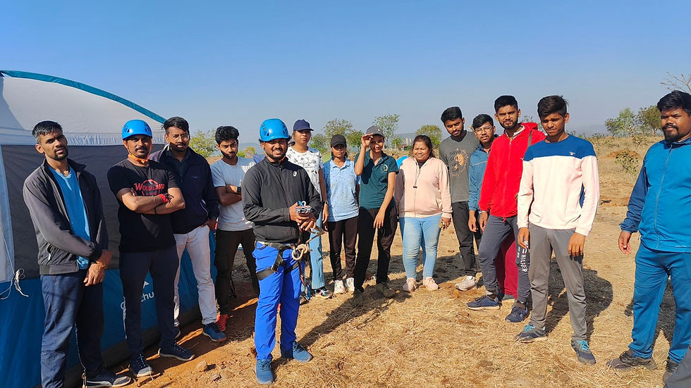 A group of people stand outdoors, one in a blue helmet with climbing gear. They're near a tent on dry grass under a clear blue sky.