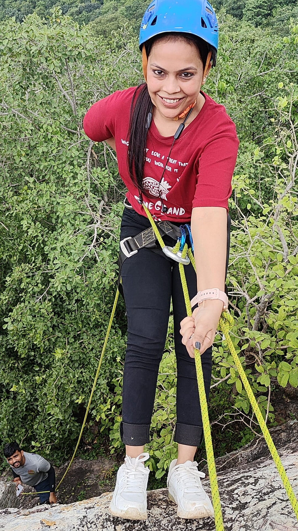 Woman in red shirt and blue helmet rock climbing with ropes, smiling. Green forest background. Man watching below.