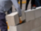 Worker in gray shirt and gloves stacking concrete blocks outdoors, with a hammer nearby. Overcast lighting and construction setting.