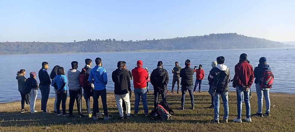 A group of people stands near a lake, listening to a speaker on a sunny day. Green hills and blue sky in the background.