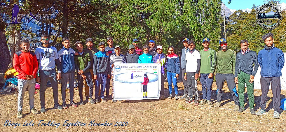 Group of hikers poses with a "Bhirga Lake Trekking Expedition 2020" sign amid a forest. They wear colorful outdoor gear, creating a vibrant scene.