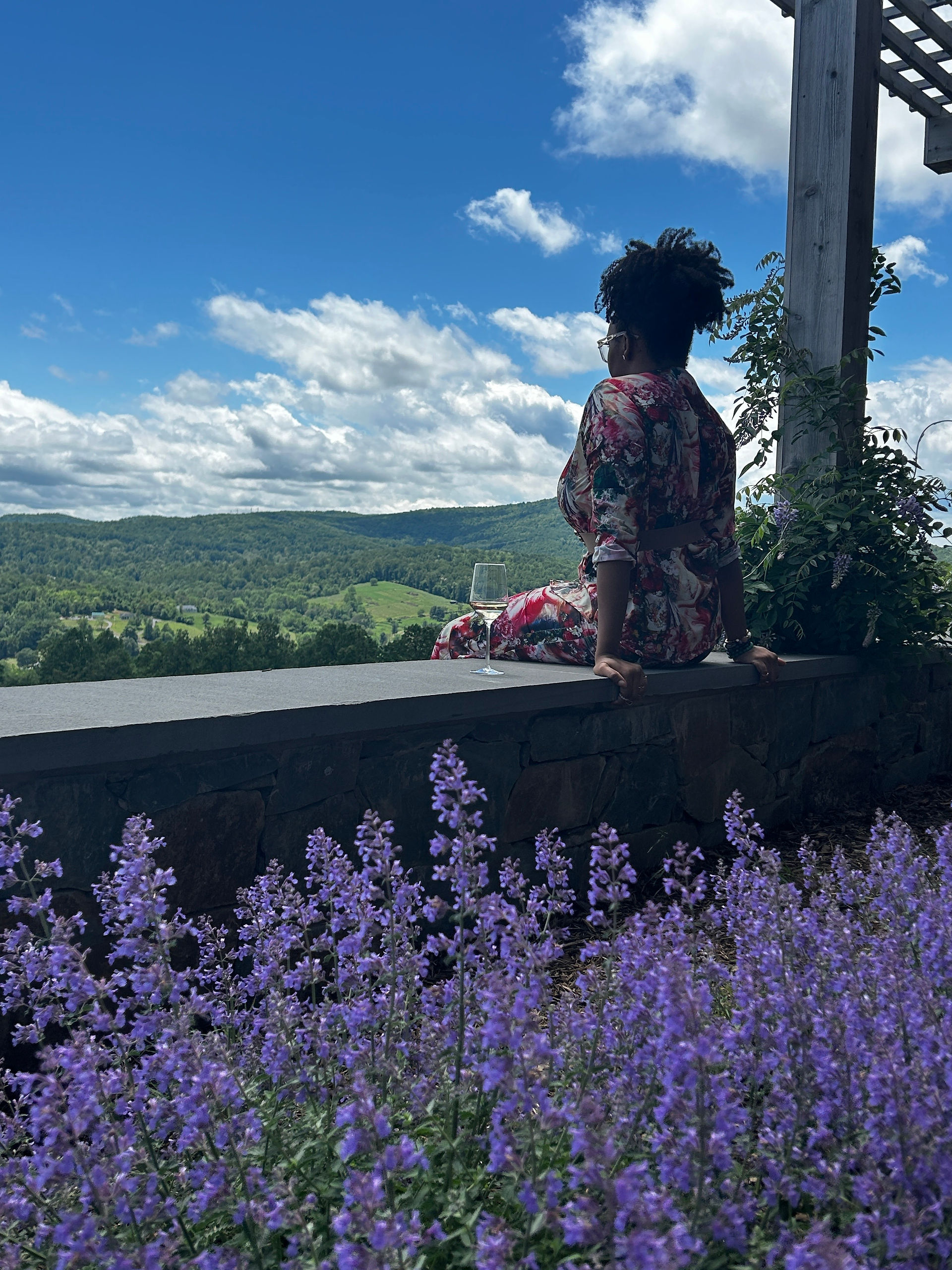 Elle sitting on a ledge overlooking a valley with back to camera and lilac flowers in foreground