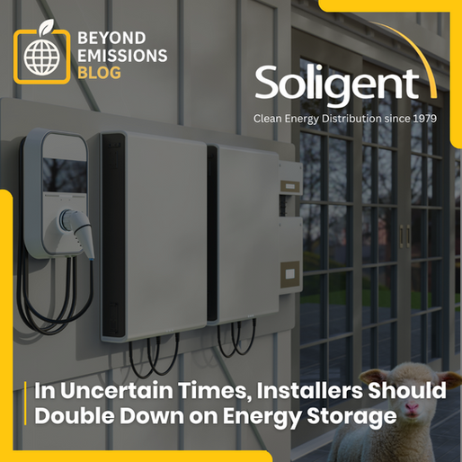 A modern home exterior features two white battery storage units mounted on the wall, next to an electric vehicle charging station. In the foreground, a small, white lamb looks towards the viewer. The image is framed with a yellow border. In the top left corner, a yellow square with a globe icon and a green leaf is displayed, along with the text "BEYOND EMISSIONS BLOG". In the top right corner, the Soligent logo is visible with the text "Clean Energy Distribution since 1979". At the bottom of the image, the text reads "In Uncertain Times, Installers Should Double Down on Energy Storage".