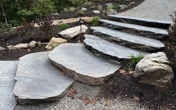 Custom granite slab steps lead from a flagstone walkway to a gravel driveway, bordered by boulders, shrubs, mulch, and a landscape light.