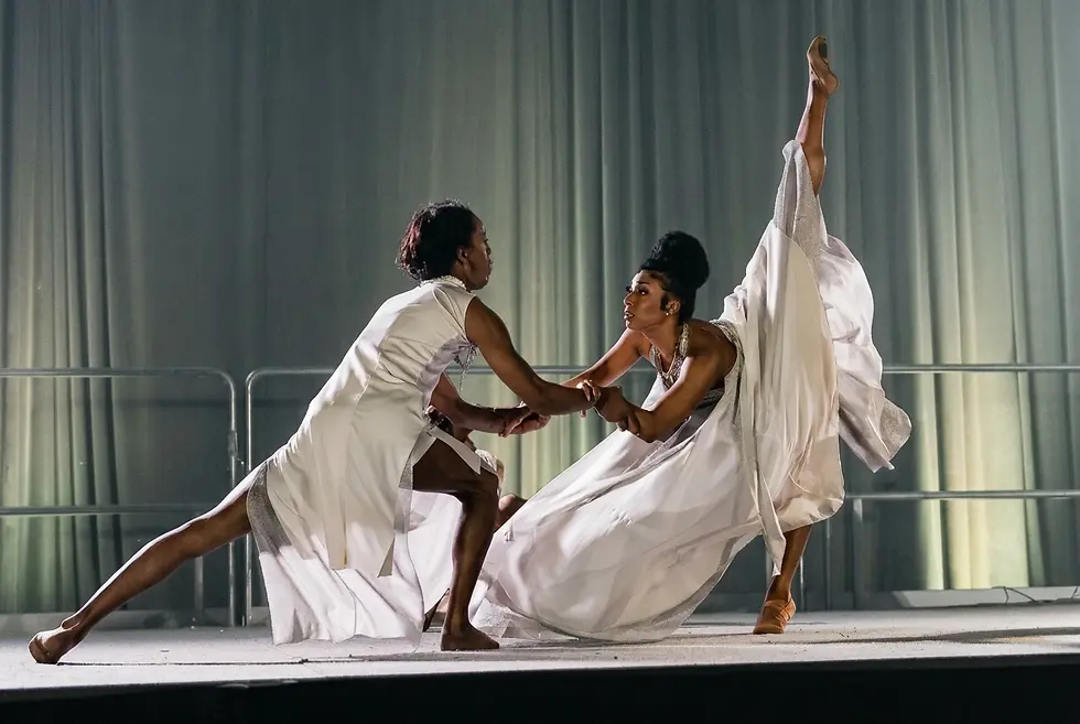 Performance photography of two Black women dancing expressively in flowing white costumes on a stage. Their dynamic poses convey a sense of dramatic artistry.