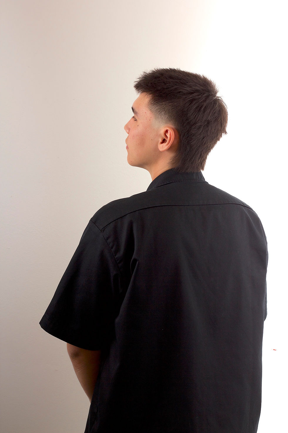 Moderní pánský střih Mullet na rovných a nepoddajných vlasech. Pohled zezadu z poloprofilu, Barber Street Praha Žižkov. Muž v černé košili.