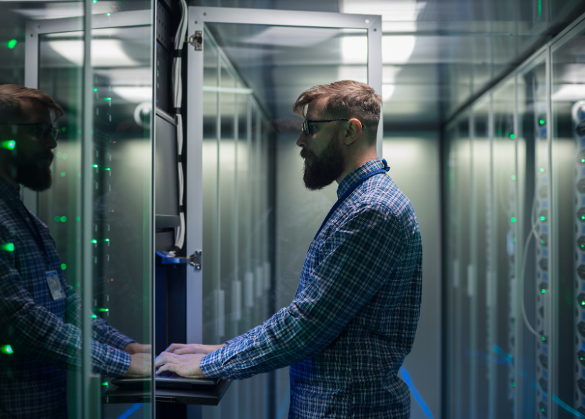 Man working on a computer in a server room, wearing glasses.