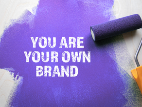 Purple paint roller with orange handle on wood, creates a bold spot reading "YOU ARE YOUR OWN BRAND" in white text. Motivational vibe.