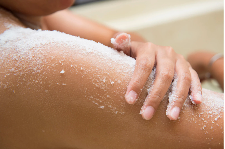 A person applies white scrub to their shoulder, showcasing exfoliation. The close-up shot emphasizes texture and a calm, self-care routine.