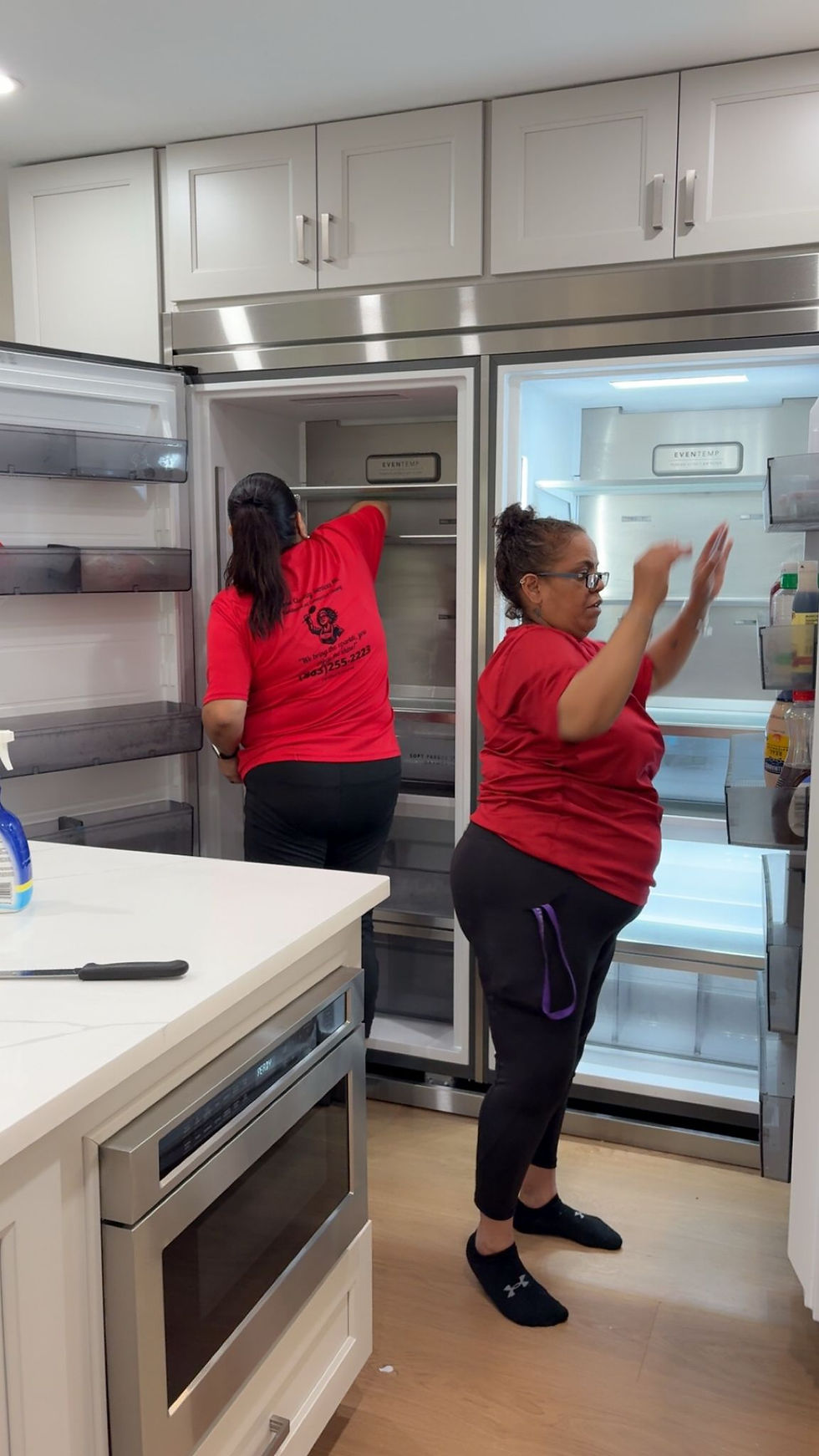 Two people in red shirts clean open refrigerators in a modern kitchen. One wipes inside while the other gestures. White cabinets and wood floor.