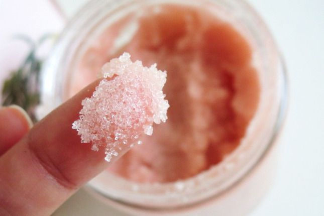 Finger with pink sugar scrub above an open jar, in a light setting, conveying a fresh and exfoliating mood.