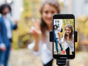 Woman flashes a peace sign while recording on a smartphone. Blurred man in blue suit in the background. Autumn leaves setting.