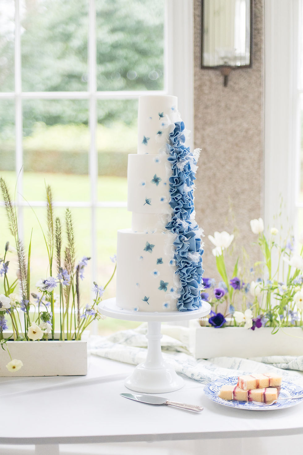 Tall white wedding cake, adorned with blue hand painted hydranges and a blue asymmetrical ruffle. It is stood upon a white cake stand next to white and blue flowers.