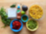Corn, edamame, roasted red peppers, cilantro, feta, jalapeƱo, avocado, lime, TajĆn seasoning, and oil on a wooden surface.