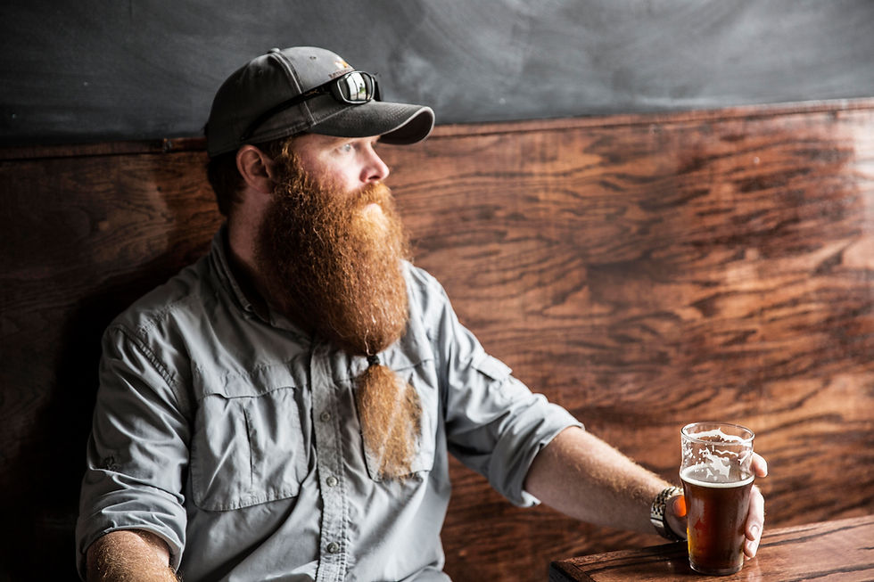 Man with a long red beard sits by a large window drinking a stout beer