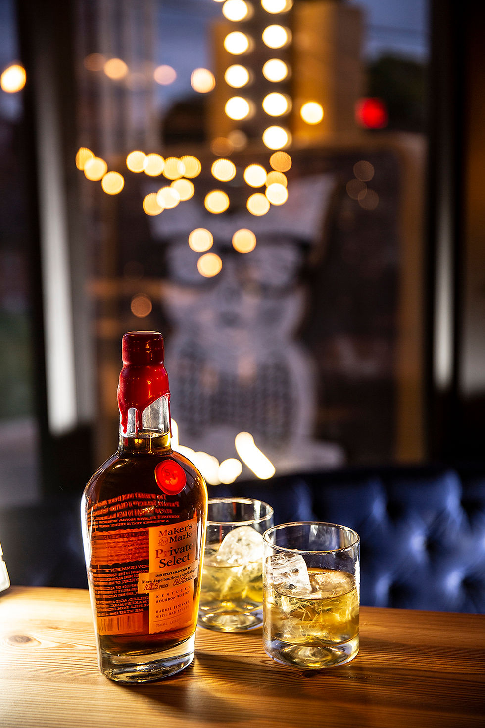 bottle of maker's mark bourbon with two glasses of bourbon on ice with twinkle lights above