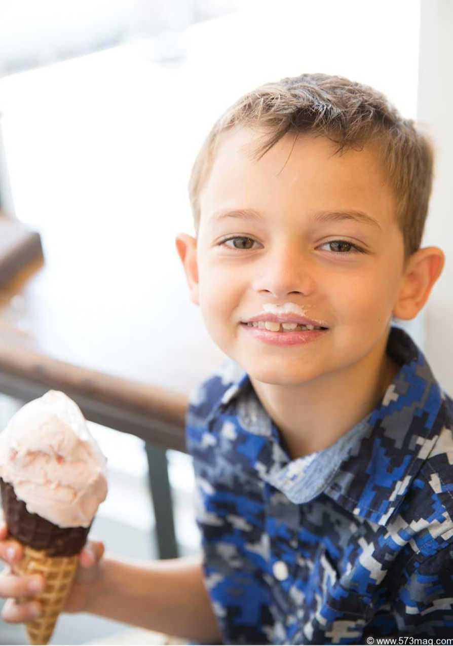 A happy child smiles at the camera with an ice cream mustache and a large ice cream waffle cone covered in chocolate dip in his hand