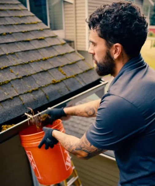 Man in gloves scooping debris from roof gutter into a red bucket during home maintenance