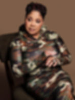 A pensive Black woman sitting on a chair wearing an Army fatigue-printed dress.