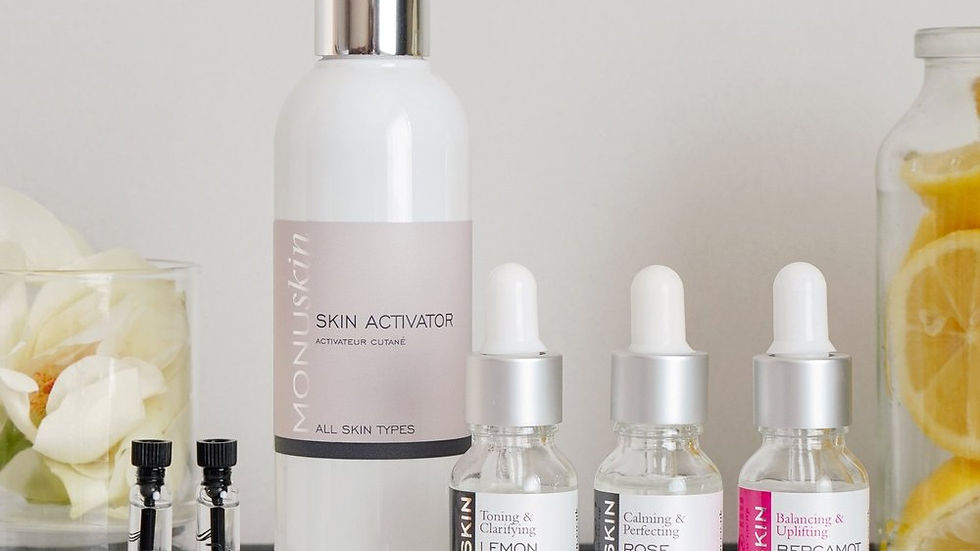 Monuskin skincare bottles and droppers on a marble surface, with flower petals and lemon slices in jars in a bright spa setting