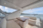 Spacious white yacht lounge deck with table and benches, overlooking calm blue water, boats, and a sunny cloudy harbor sky.