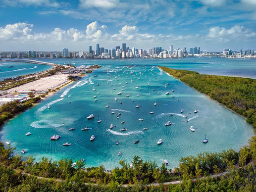 Aerial view of numerous boats in a turquoise miami bay, surrounded by green land and a city skyline under a cloudy blue sky.
