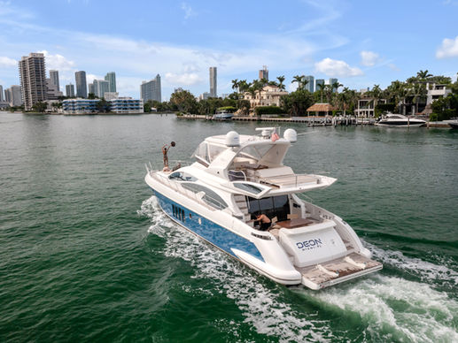 Luxury yacht DEON Miami FL cruising past waterfront homes and skyline, with a person waving on deck under a bright blue sky
